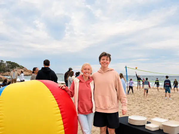 Two college students wearing sweatshirts stand on the beach next to a large inflatable beach ball