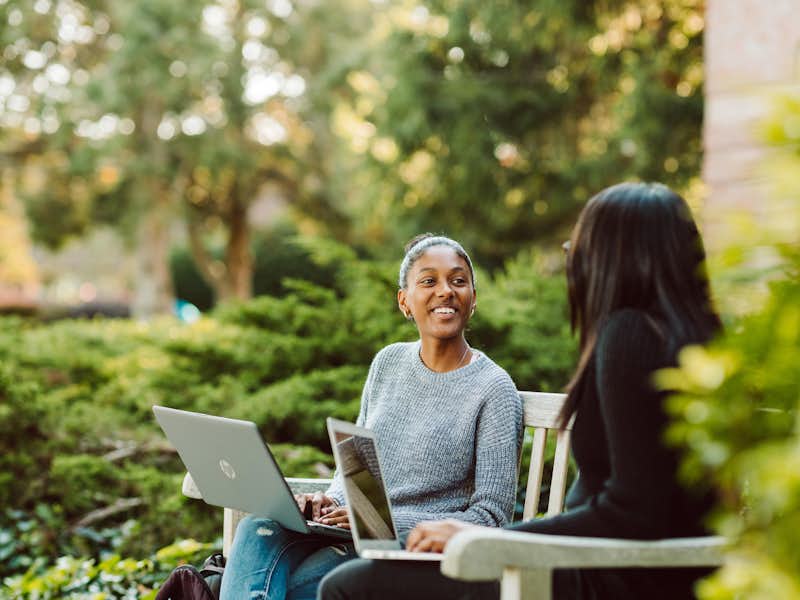 Two students work on their laptops outdoors while chatting.