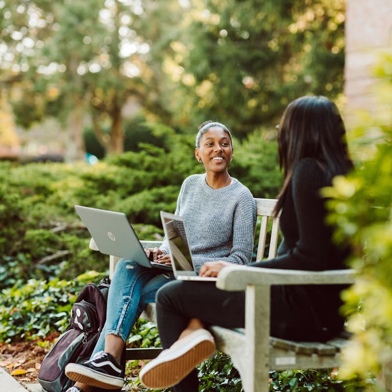 Two students work on their laptops outdoors while chatting.
