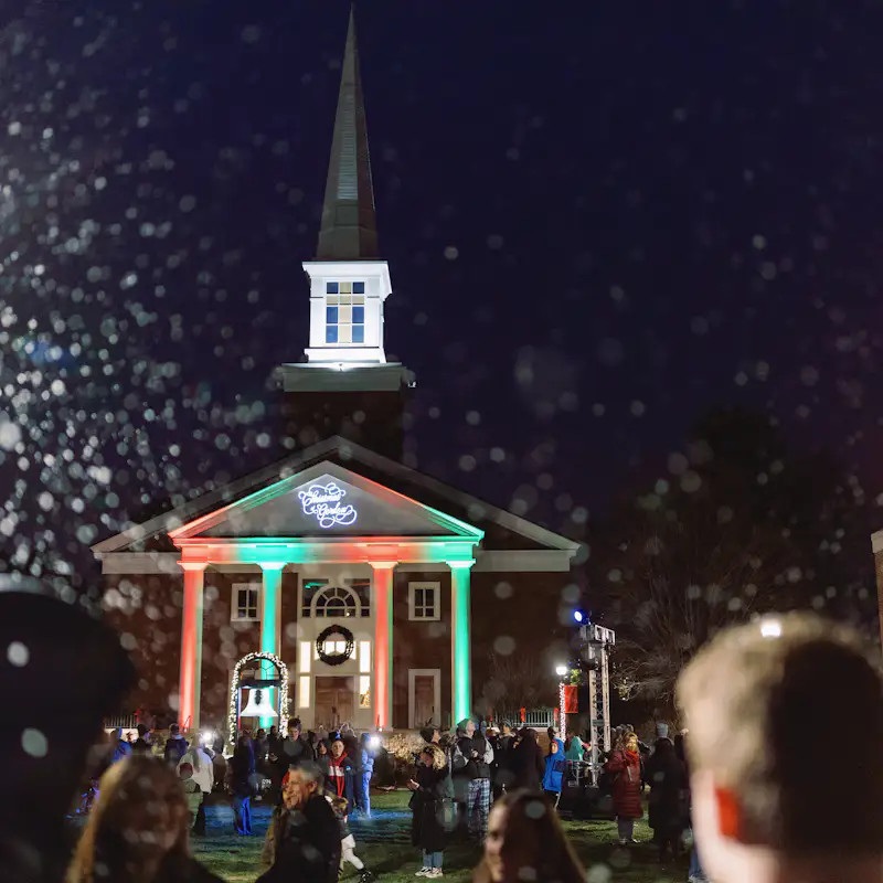 The A. J. Gordon Memorial Chapel is lit up by green and red colored lights. A crowd of people stand outside as it snows.