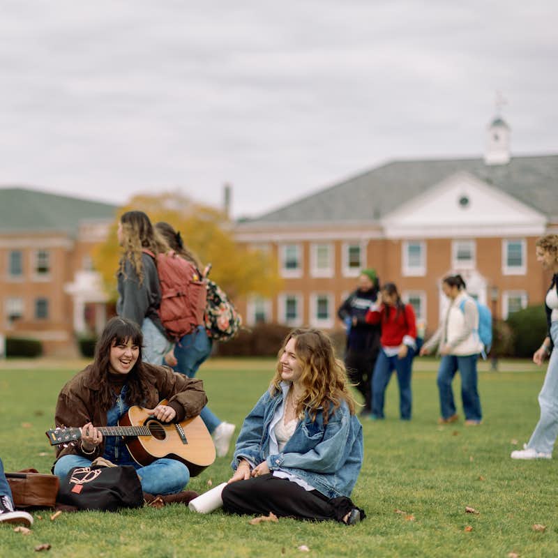 Students sit together on the quad, playing guitar and laughing while other students walk to class
