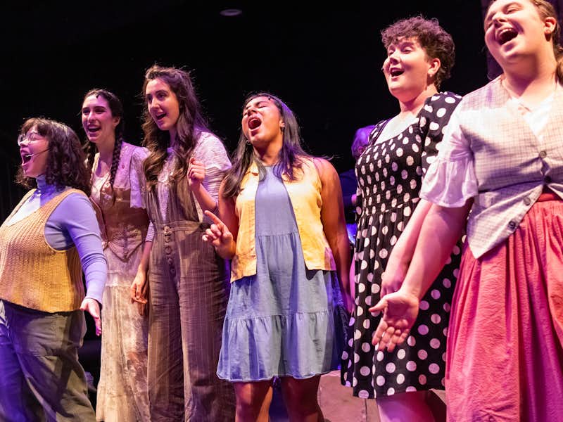 Six students wearing colorful outfits sing passionately during a performance of Godspell