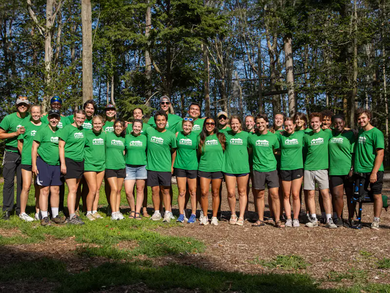 A group of teenagers stand with their arms around each other in the forest, they are all wearing matching green shirts