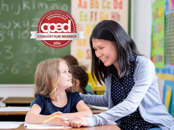 A teacher with dark hair and a light blue cardigan works one on one with an elementary school student at her desk. Text reads: CPED Consortium Member