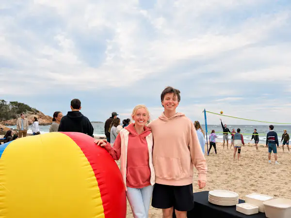 Two college students wearing sweatshirts stand on the beach next to a large inflatable beach ball