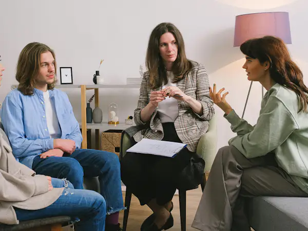 A group of adults sit on couches and chairs in a circle, talking to a therapist who is taking notes