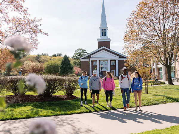 A group of students walk on grass in the sun in front of the A. J. Gordon Memorial Chapel