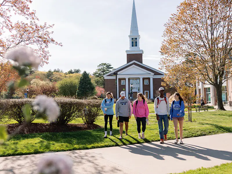 A group of students walk on grass in the sun in front of the A. J. Gordon Memorial Chapel