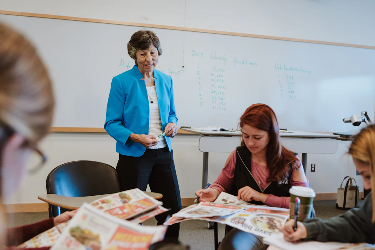 A teacher wearing a blue blazer leans over and talks with college students.