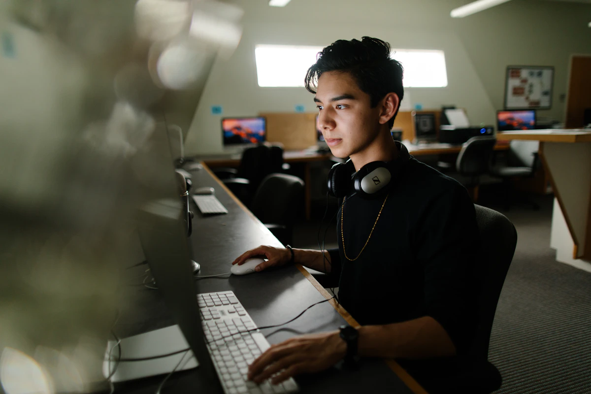 A student with headphones around his neck works on editing a video on a desktop computer.