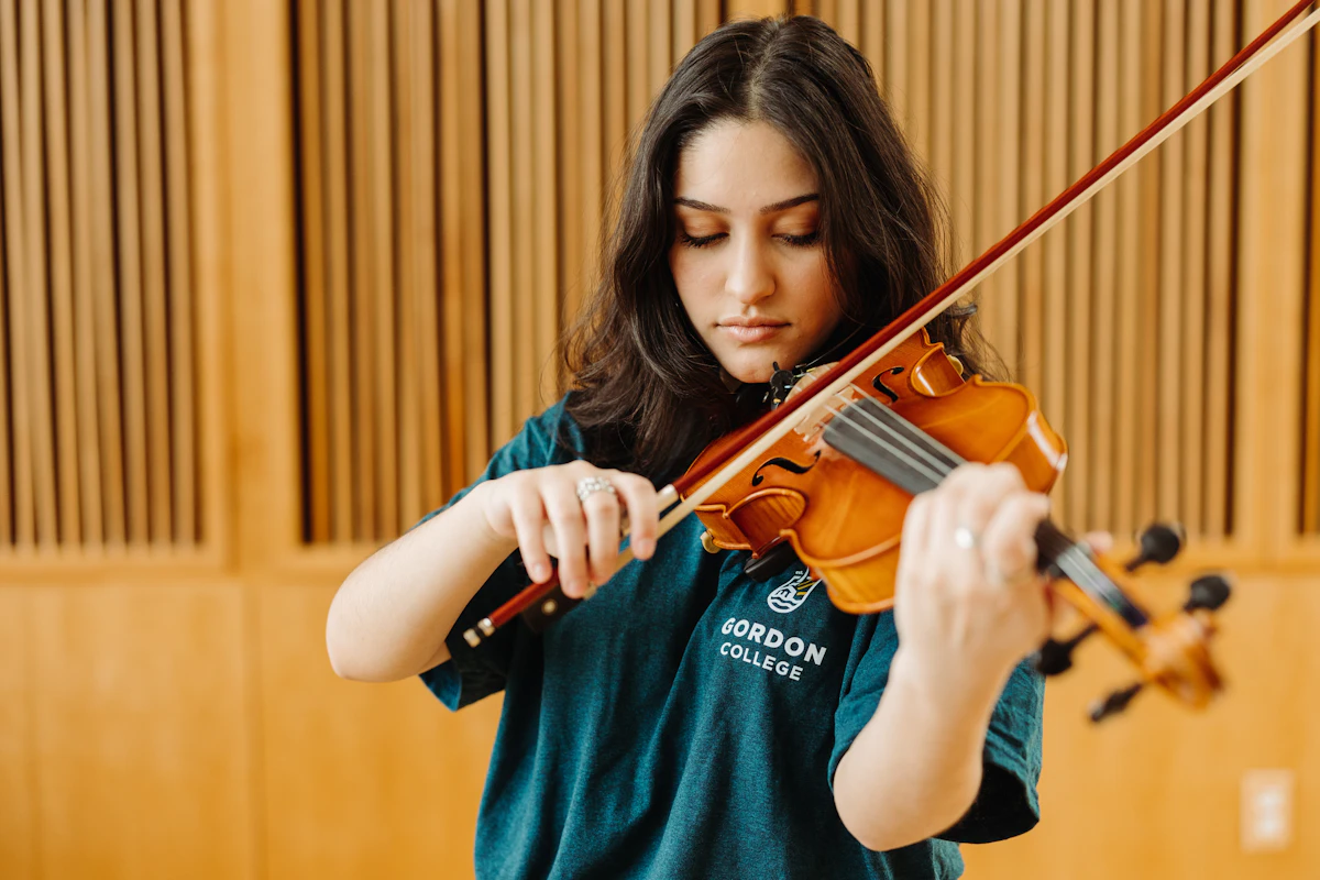 A young woman plays a violin. She has dark hair and wears a navy blue shirt with the Gordon College logo on it.