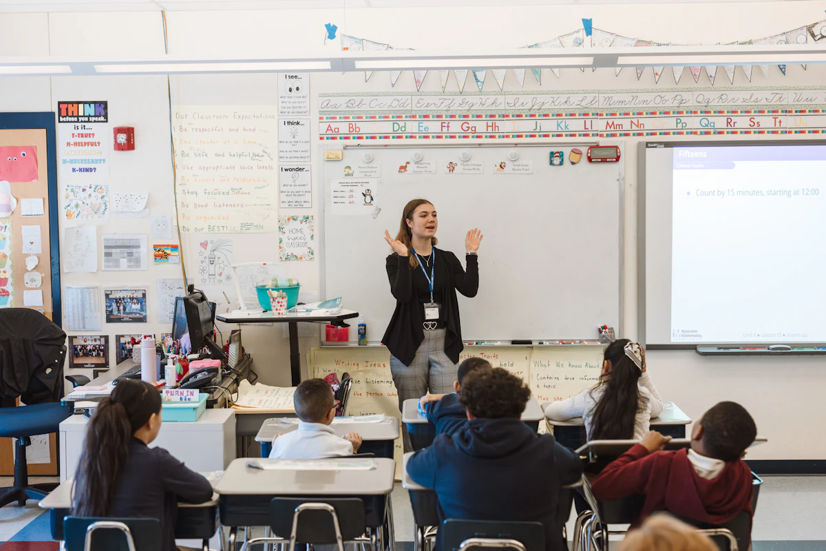 A teacher stands at the front of an elementary school classroom and claps her hands.