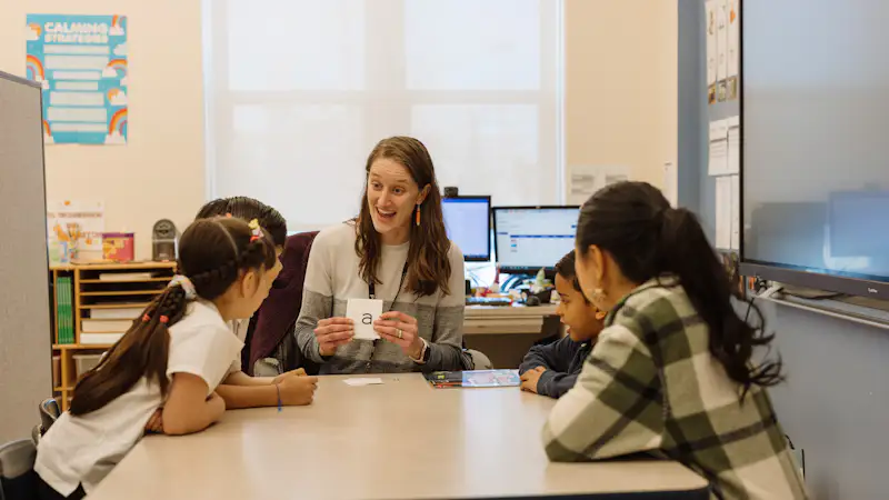 A teacher works with four elementary students at a table. She holds a card with the letter a in her hands.