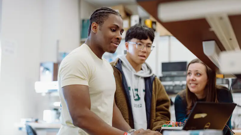 Two students work their professor on an engineering project in a lab. There is a computer, wires, and other technology on the table.