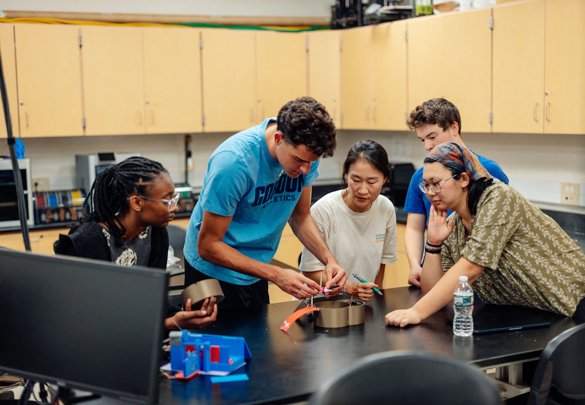 A group of college students work on a project with their professor. The group gathers in an engineering lab and lean over a table to observe a mechanical engineering project.