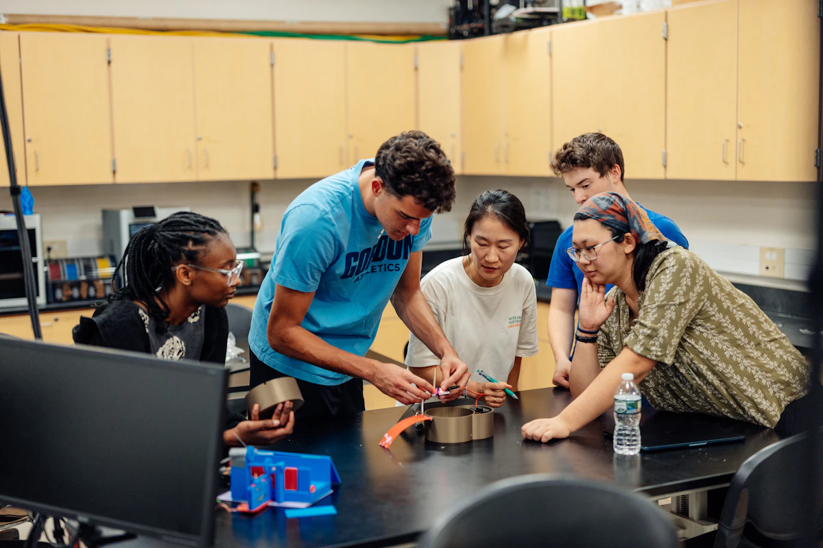 A group of college students huddle around an engineering project on a table in a lab. They work with wires and metal as their professor watches.