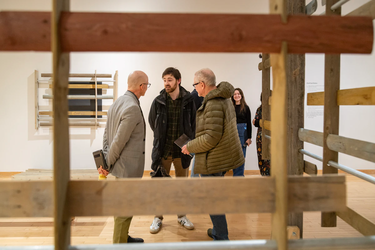 People in suits stand in the center of an art gallery, surrounded by a 3-dimensional installation made of fence posts.