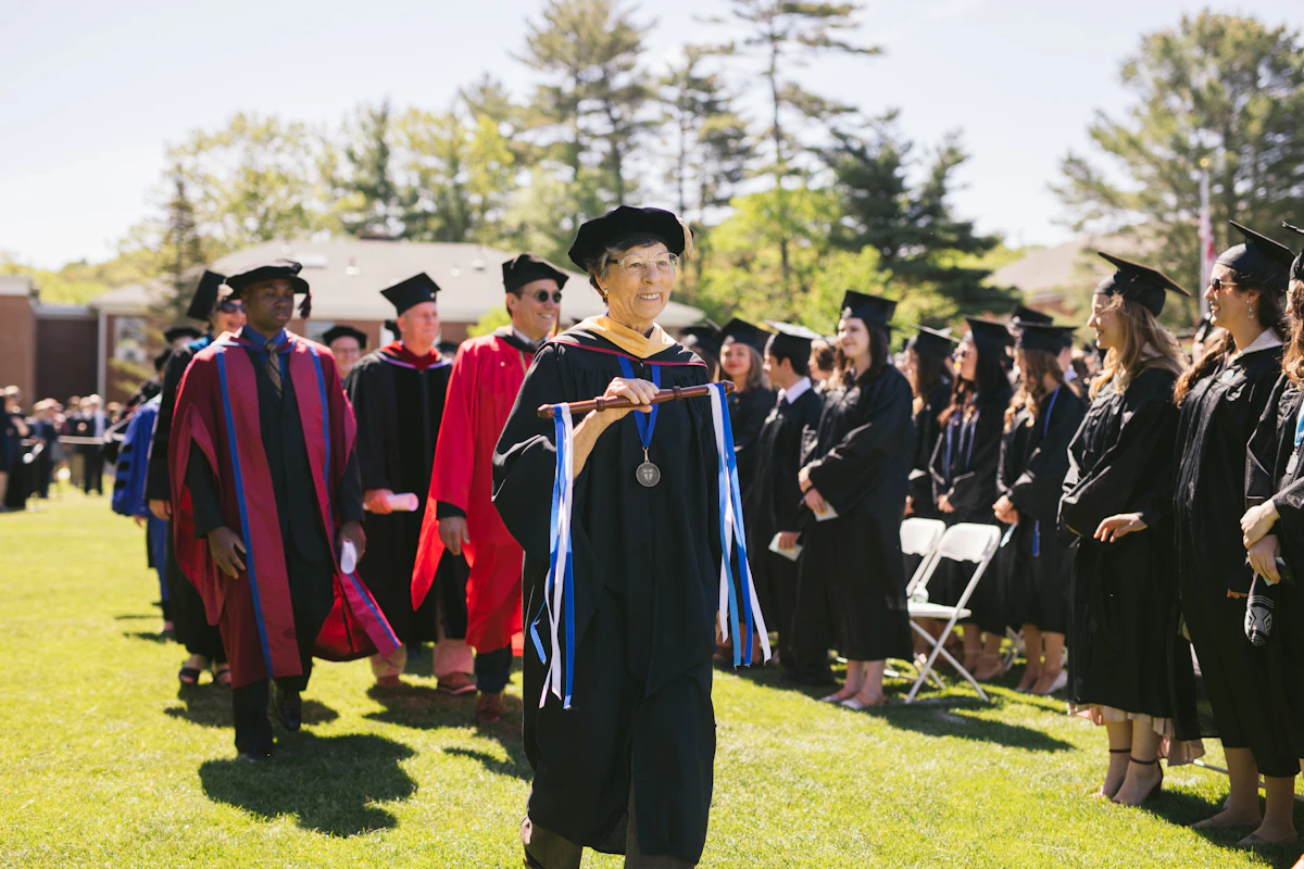 A professor holding a wooden dowel walks down a grass path at a college graduation. She wears a black cap and gown.