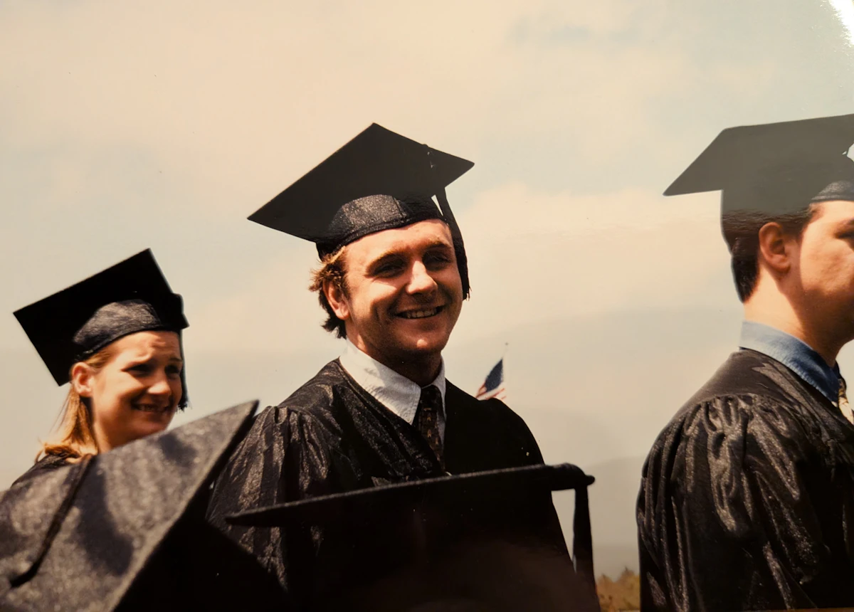 A college graduate wears a cap and gown and smiles as he walks by.