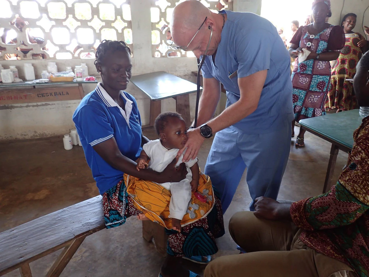 A man in blue scrubs listens to the heartbeat of a young child sitting an a woman's lap.