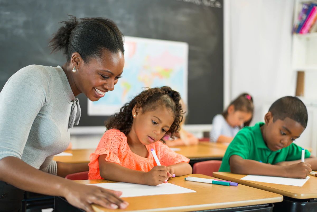 A teacher works with two elementary school students as they fill out worksheets at their desks.